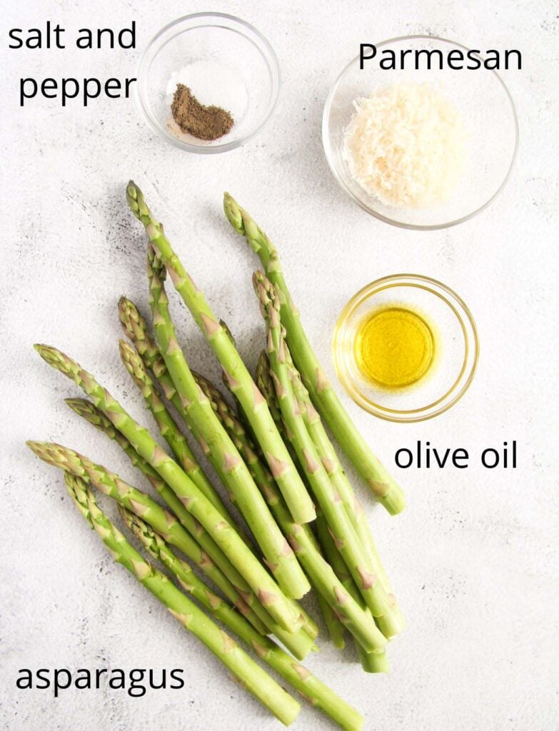 bunch of asparagus, salt, pepper, olive oil and grated parmesan in small bowls on a white table.