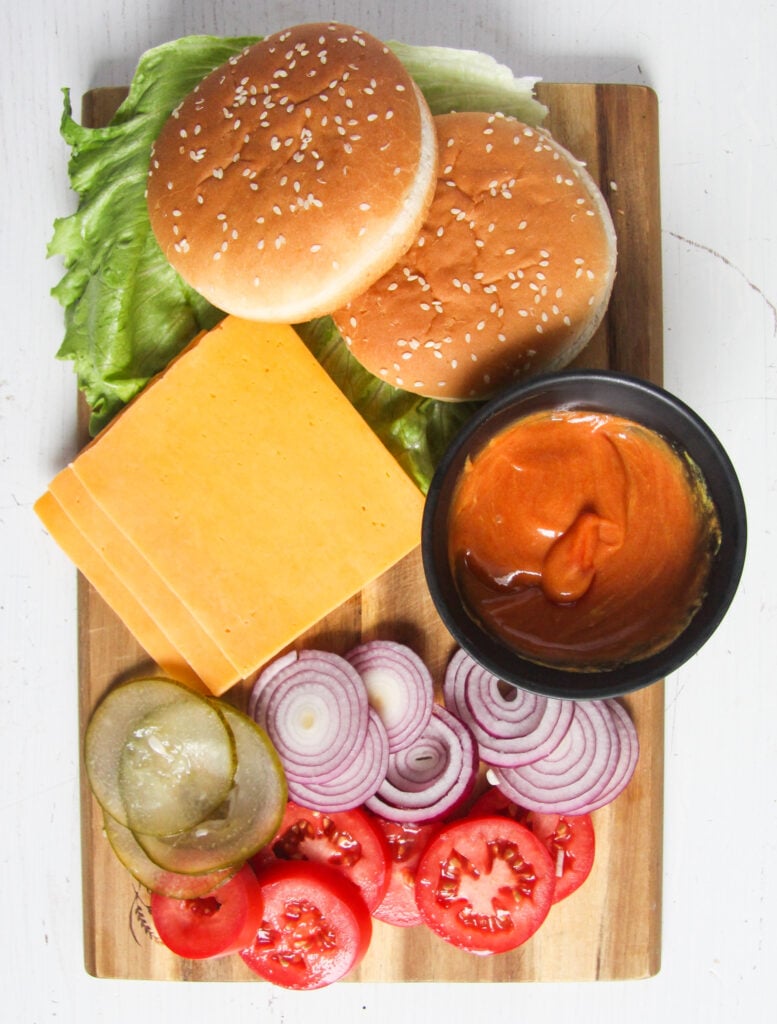 two sesame buns, cheddar slices, ketchup sauce, sliced onions, tomatoes. sour cucumbers and lettuce on a wooden board.