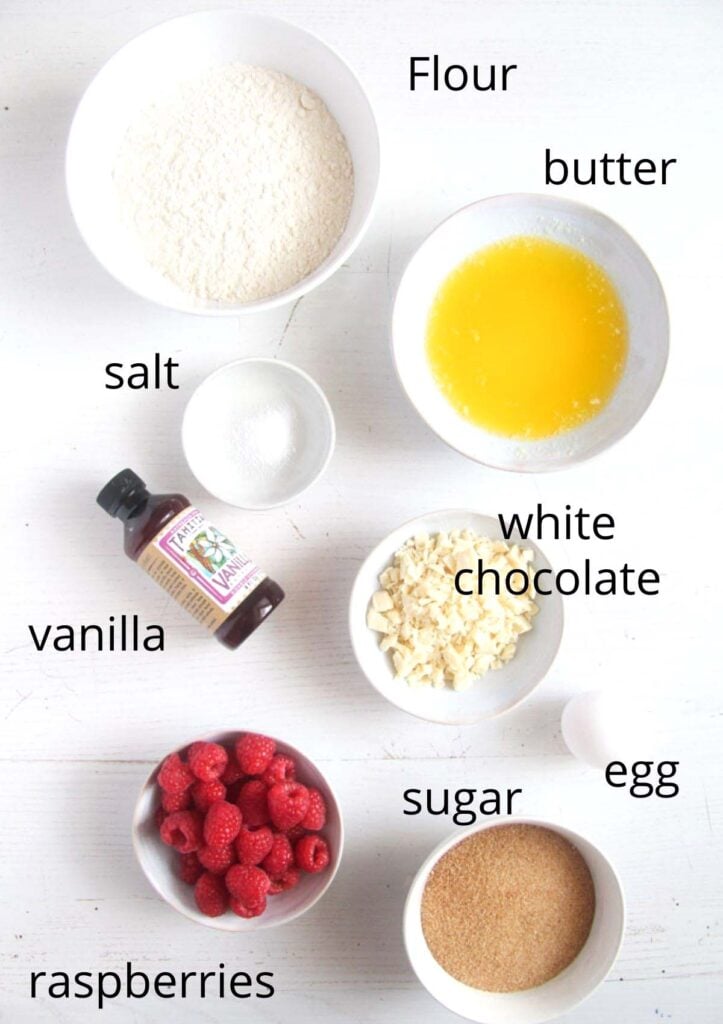 bowls with flour, melted butter, sugar, vanilla, white chocolate, egg, raspberries overhead view.