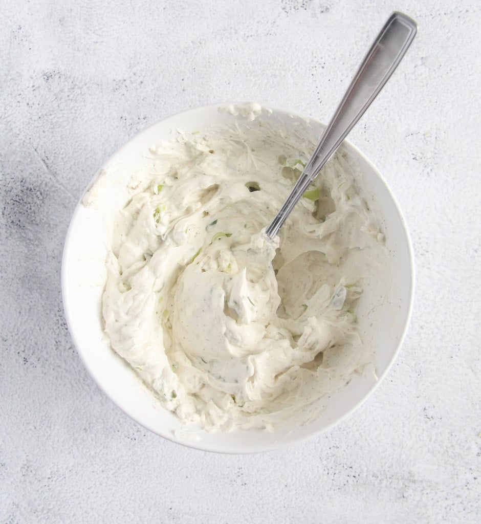 bowl of cream cheese mixed with chopped green onions and basil with a spoon in it.