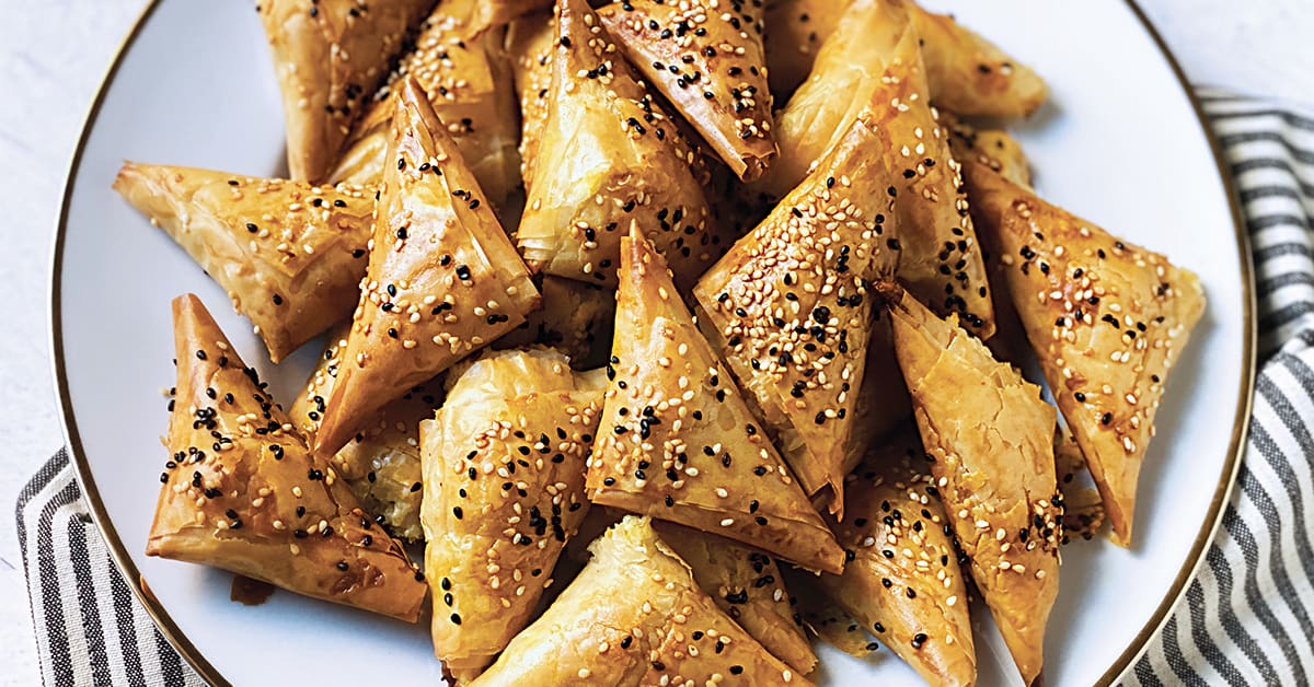 many tiropitakia cheese triangles sprinkled with nigella seeds on a white plate.