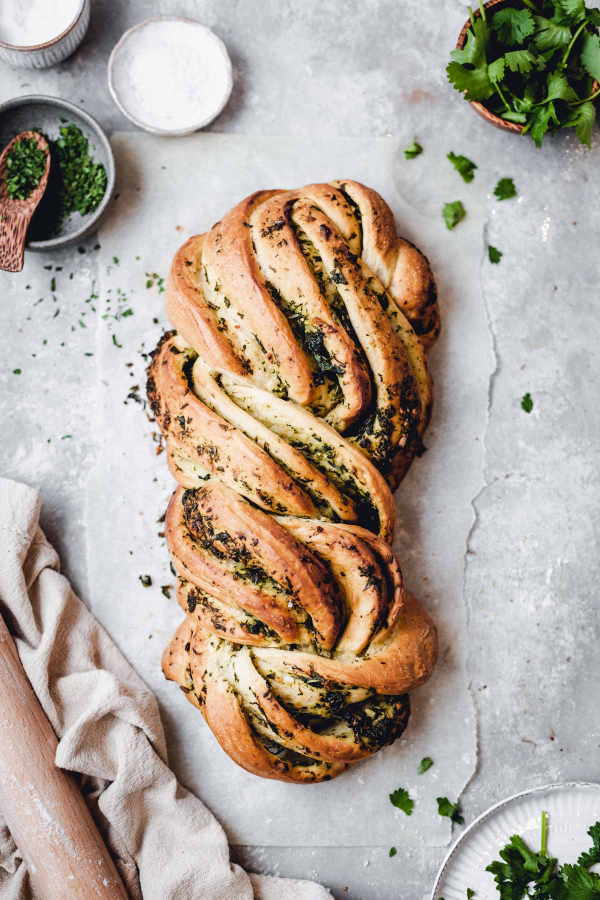 beautifully plaited vegan garlic bread on a grey table with herbs and bowls around it.