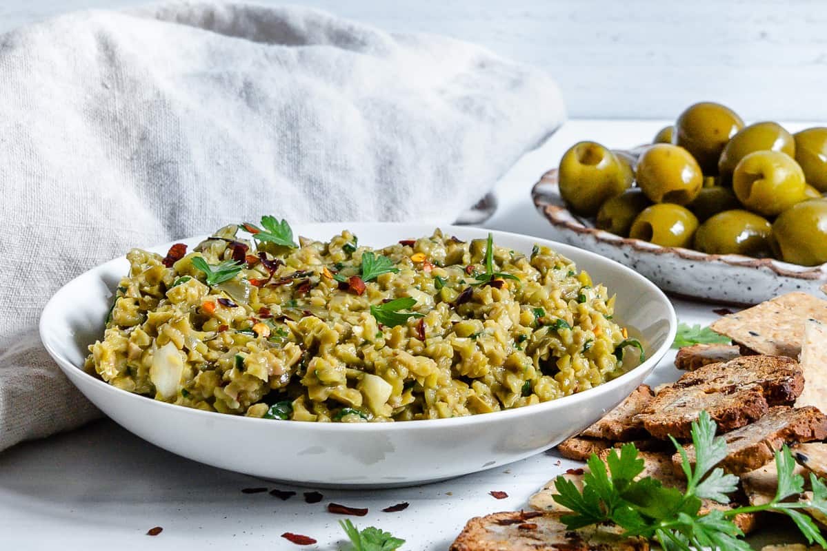 shallow bowl full with vegan green olive tapenade, many green olives in a bowl behind.