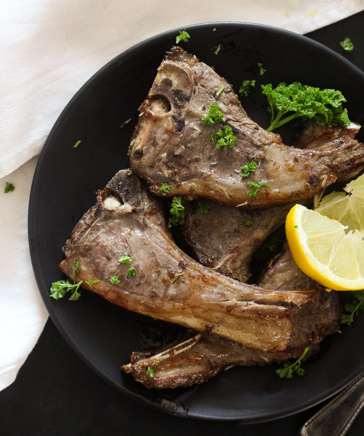 many lamb chops with lemon wedges and parsley on a black plate.