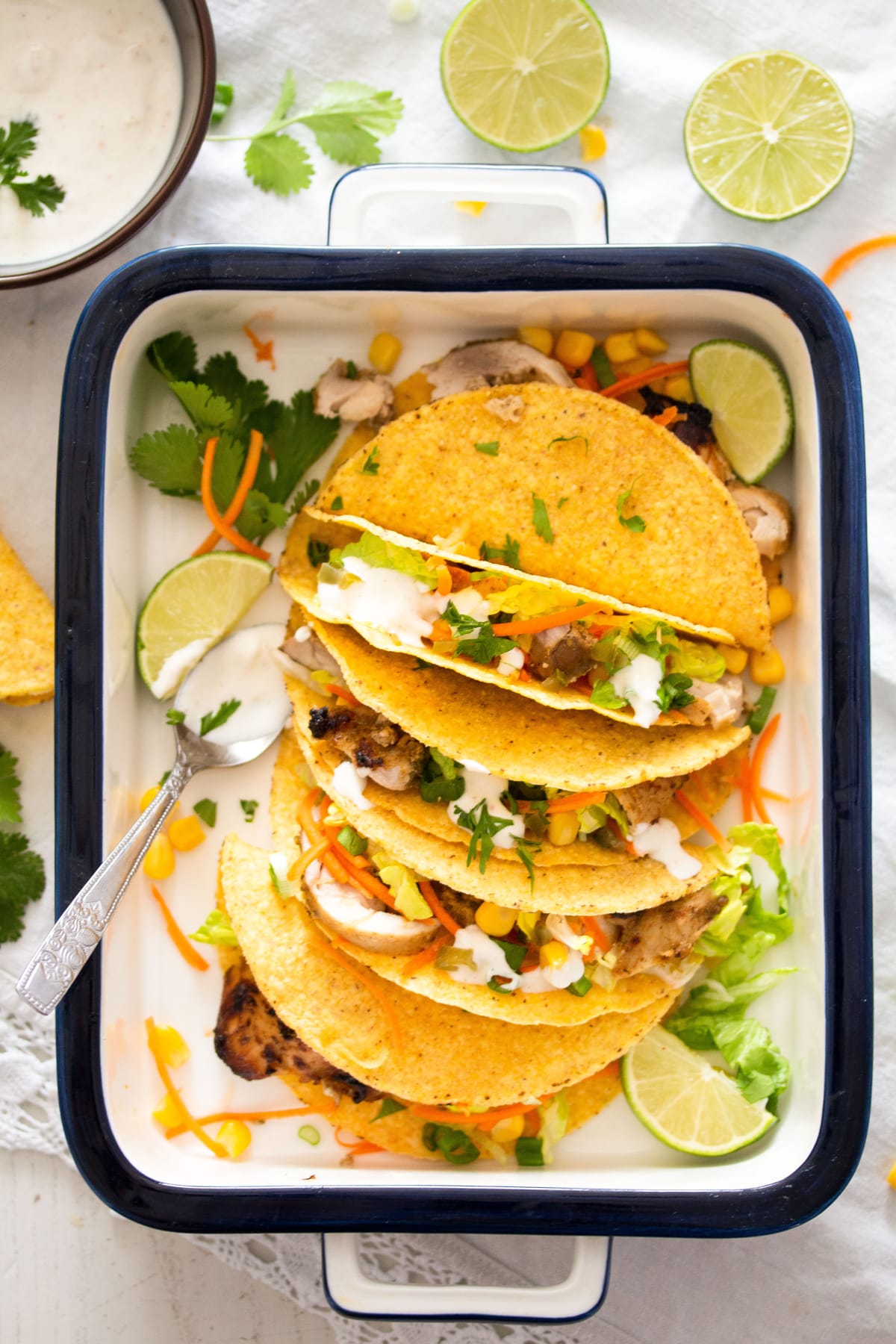 four chicken cilantro lime tacos in crunchy shell in a serving dish with limes and sauce beside it.