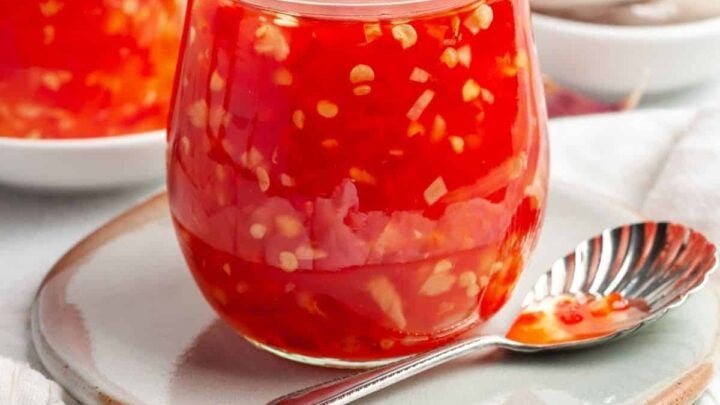 small jar full of glossy red sweet chili sauce on a small plate with a spoon.