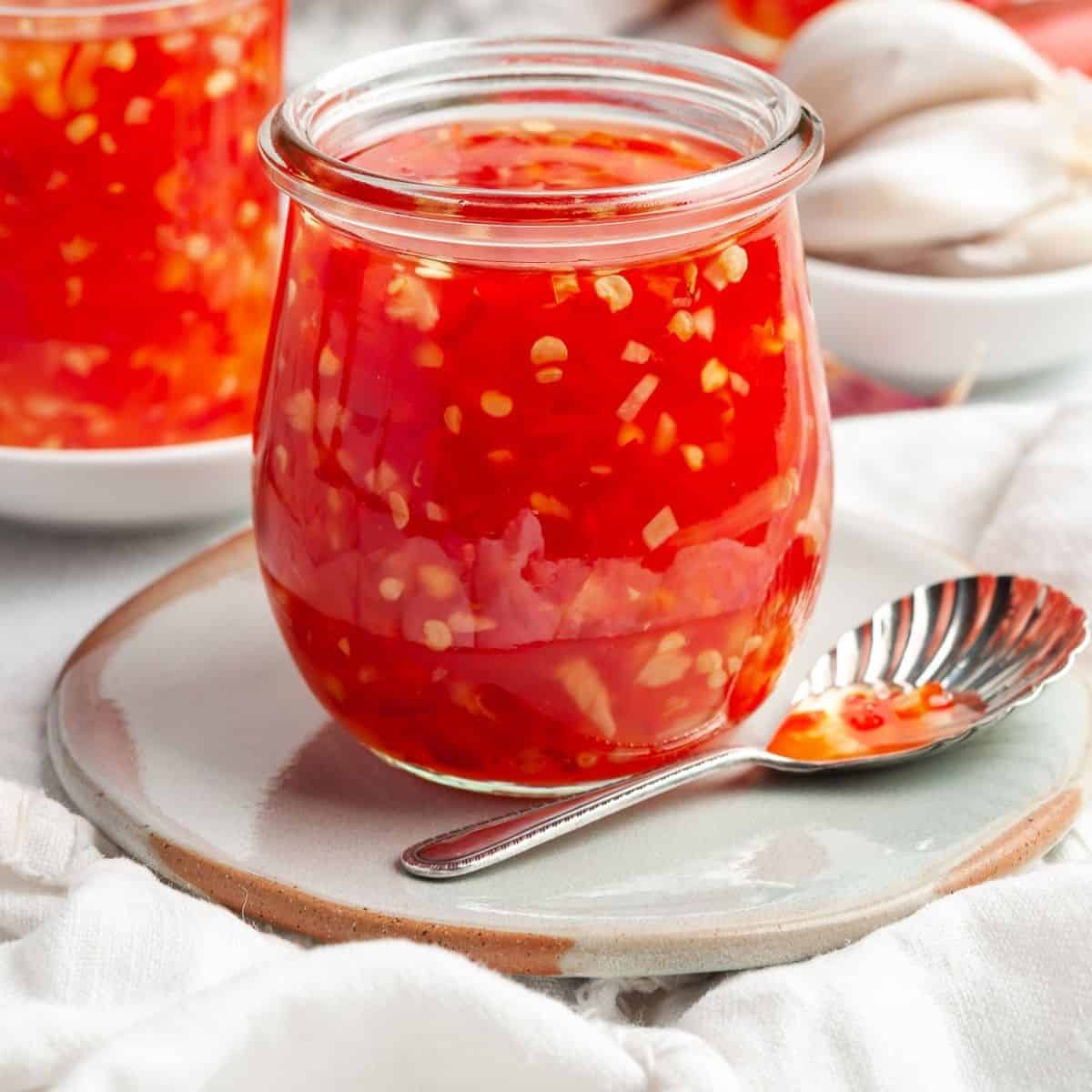 small jar full of glossy red sweet chili sauce on a small plate with a spoon.