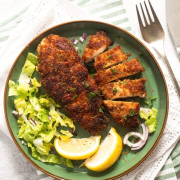 two breaded chicken breasts with lemon and salad on a green plate.