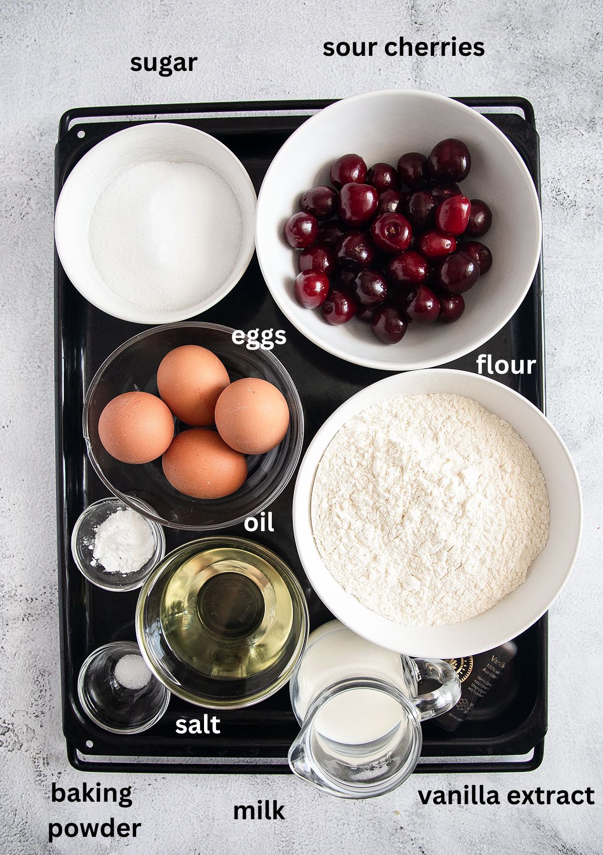 listed ingredients for cherry cake placed in bowls on a large baking tray.