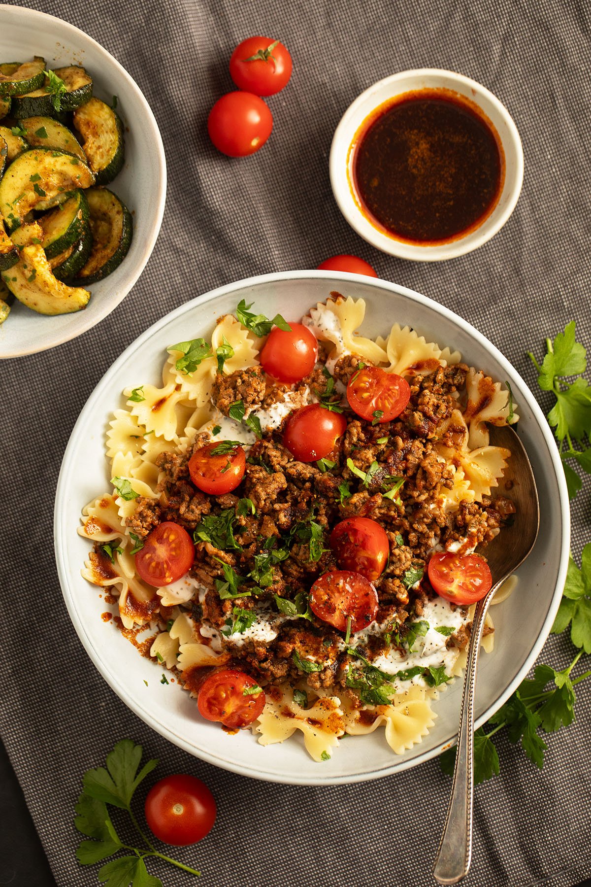 turkish pasta dish with ground beef, yogurt and halved cherry tomatoes, bowls of paprika butter and fried zucchini beside it.