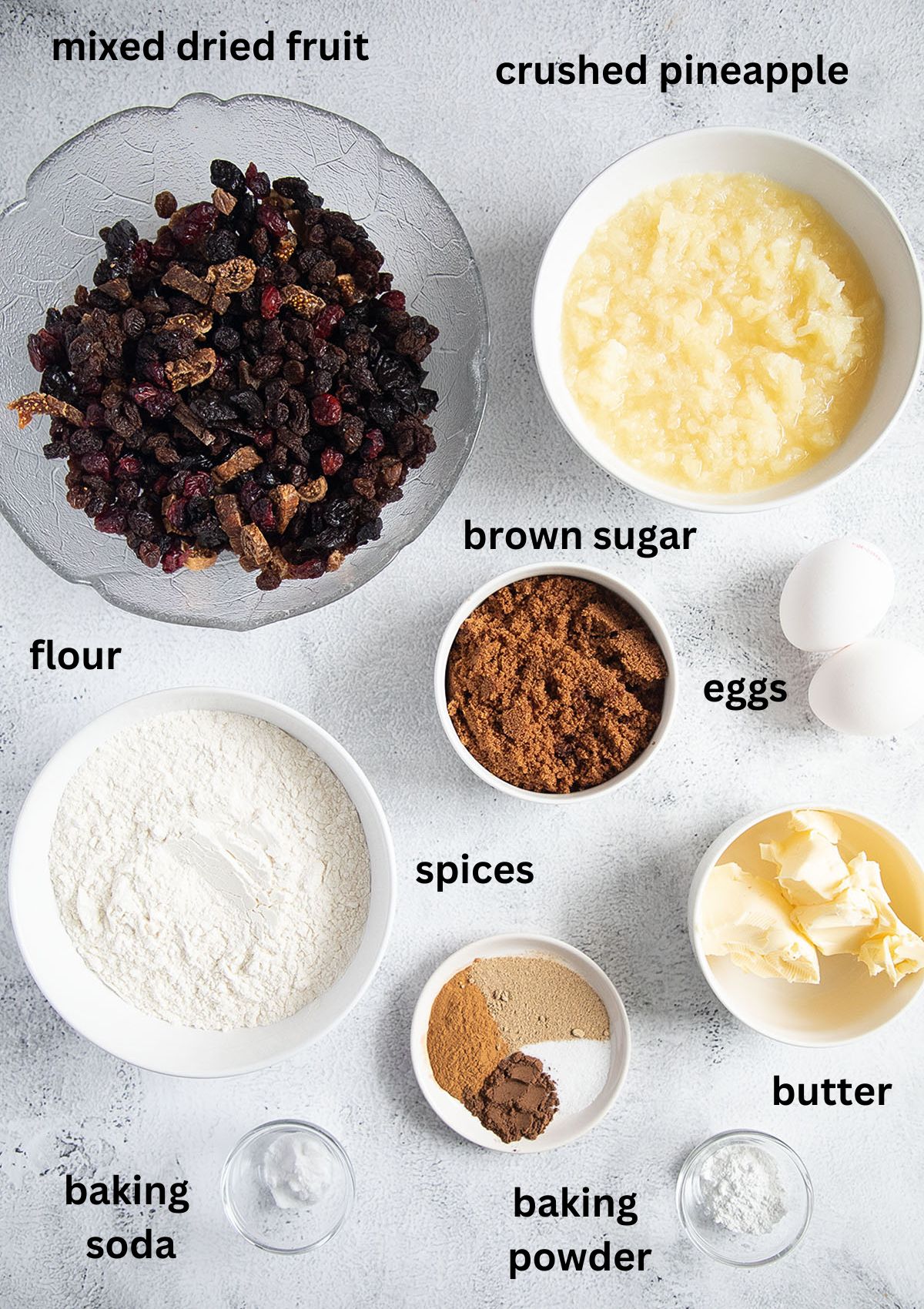 labeled ingredients for making fruit cake with canned pineapple, brown sugar, dried fruit, butter and spices.