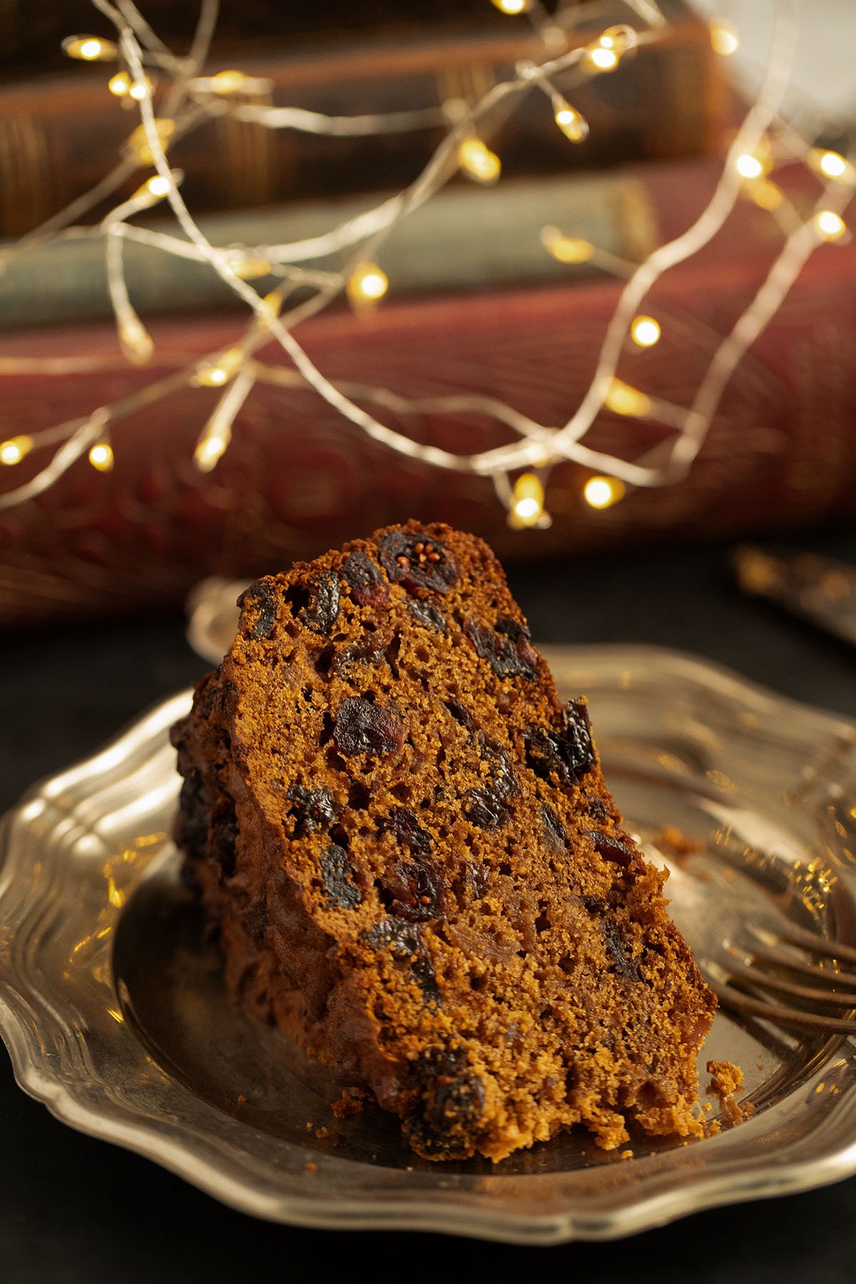 slice of pineapple fruitcake on a plate, books and christmas lights behind it.