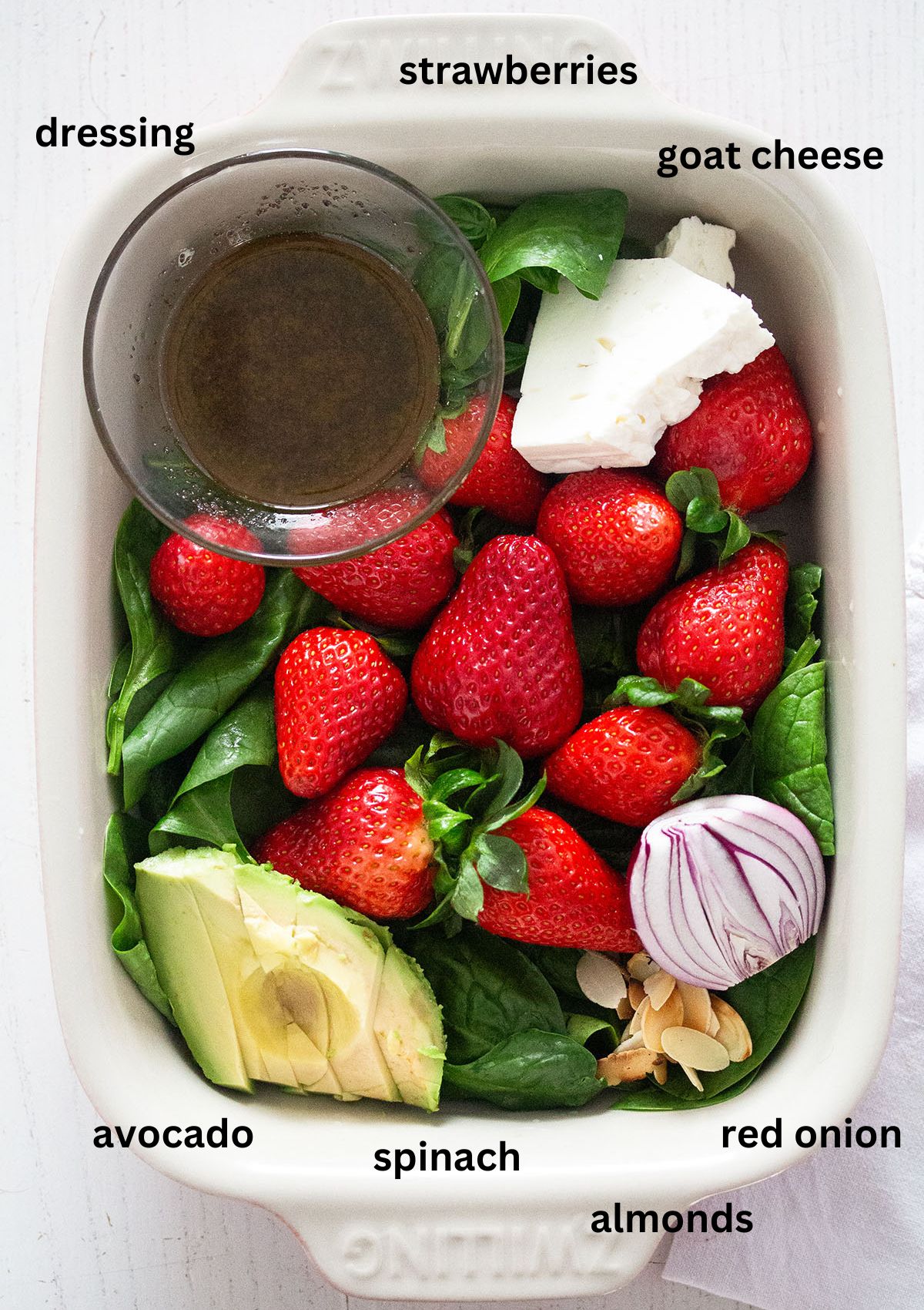 labeled ingredients for making salad with strawberries, goat cheese, spinach, dressing and avocado in a baking dish.