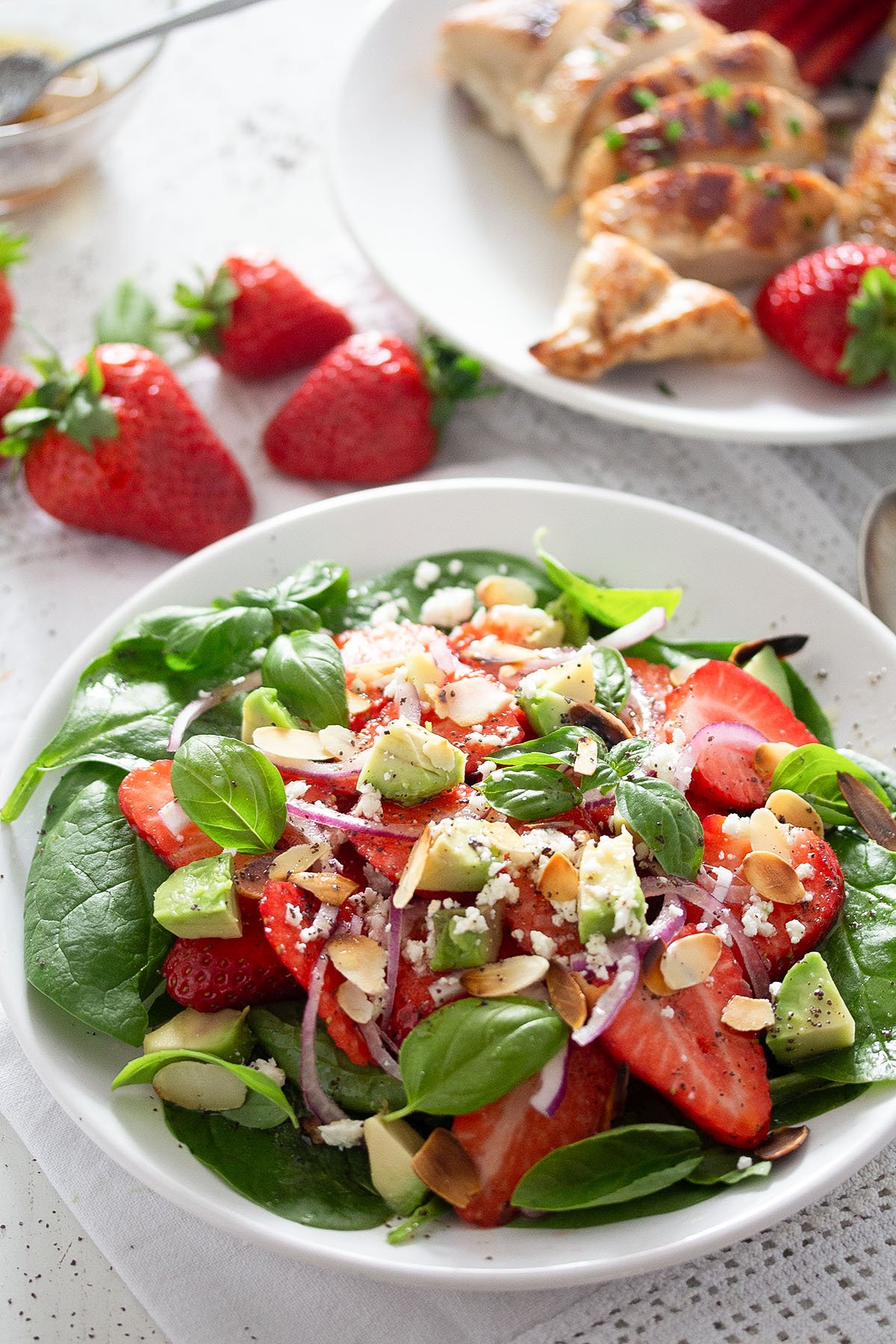 salad with spinach leaves, strawberry, goat cheese and avocado, some strawberries and grilled chicken behind.