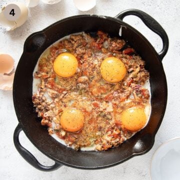 four cracked, uncooked eggs on top of sardines and tomatoes in a skillet.