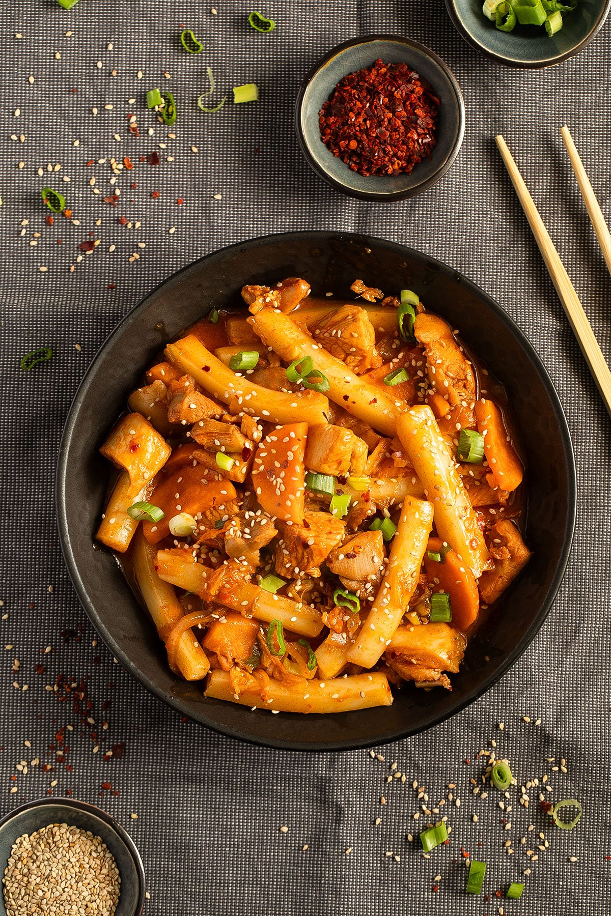 one bowl with chicken and rice cake stir fry in a bowl, small bowls with gochugaru and sesame seeds beside it.