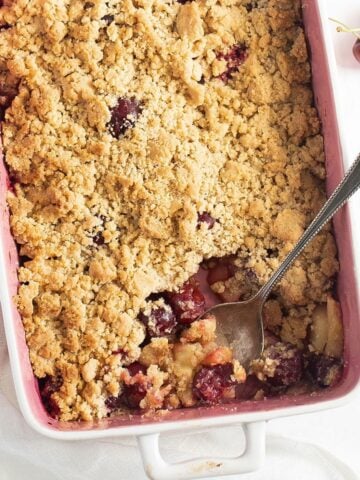 apple cherry crumble in a baking dish with a spoon in it, and a few fresh cherries around it.