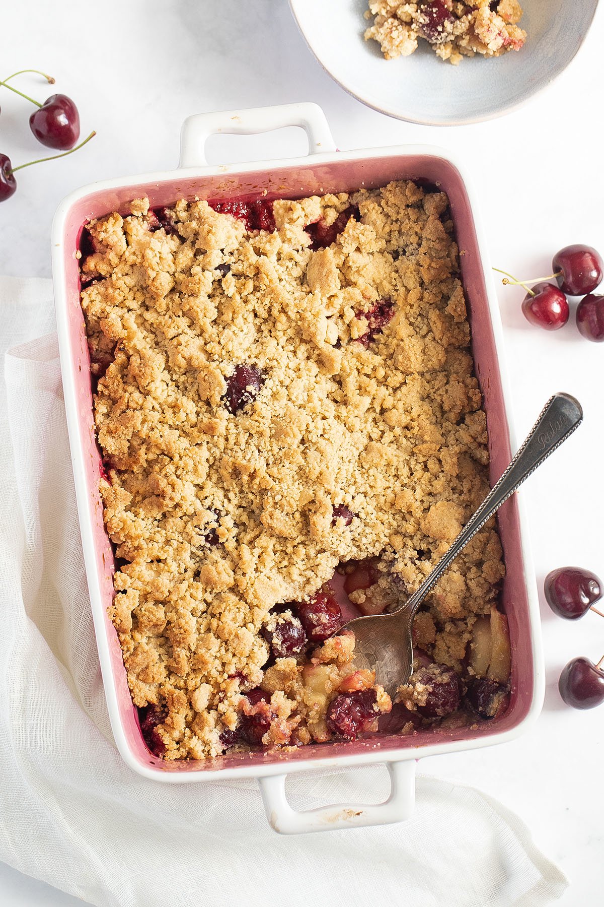 a baking dish with golden brown cherry and apple crumble and a few cherries around the dish.