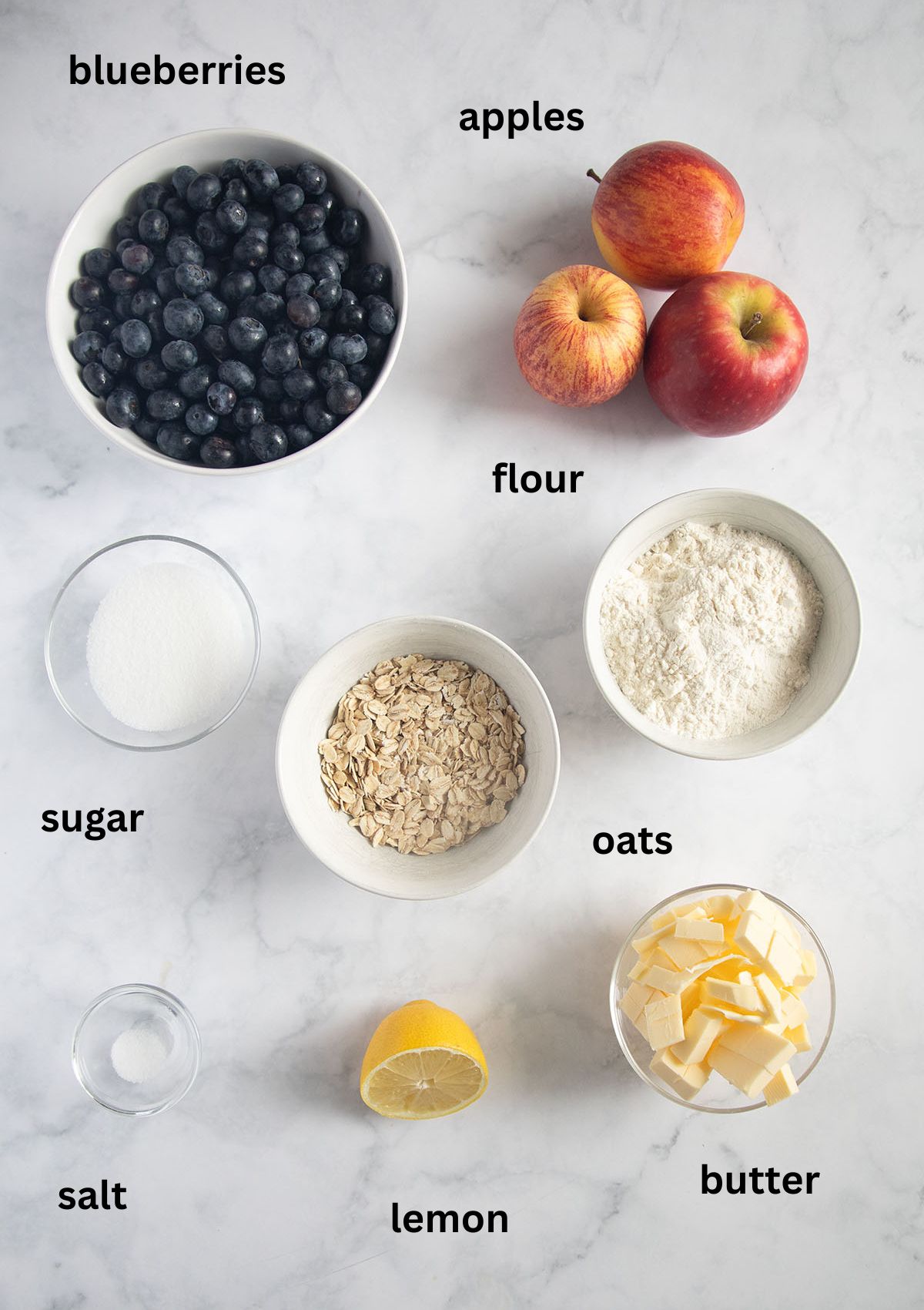 Labeled ingredients for apple and blueberry crumble recipe — apples, blueberries, rolled oats, flour, sugar, and butter in bowls on a wooden counter.