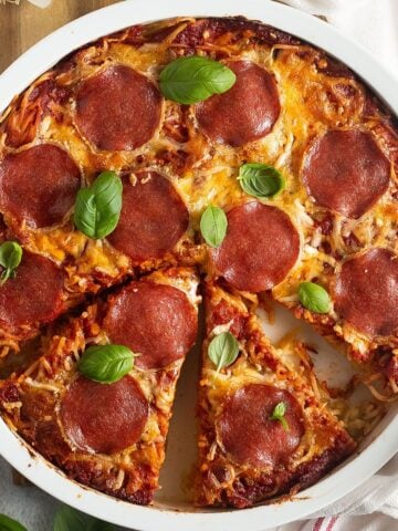 Golden baked spaghetti pizza with melted cheese and salami slices, served in a round ceramic dish.