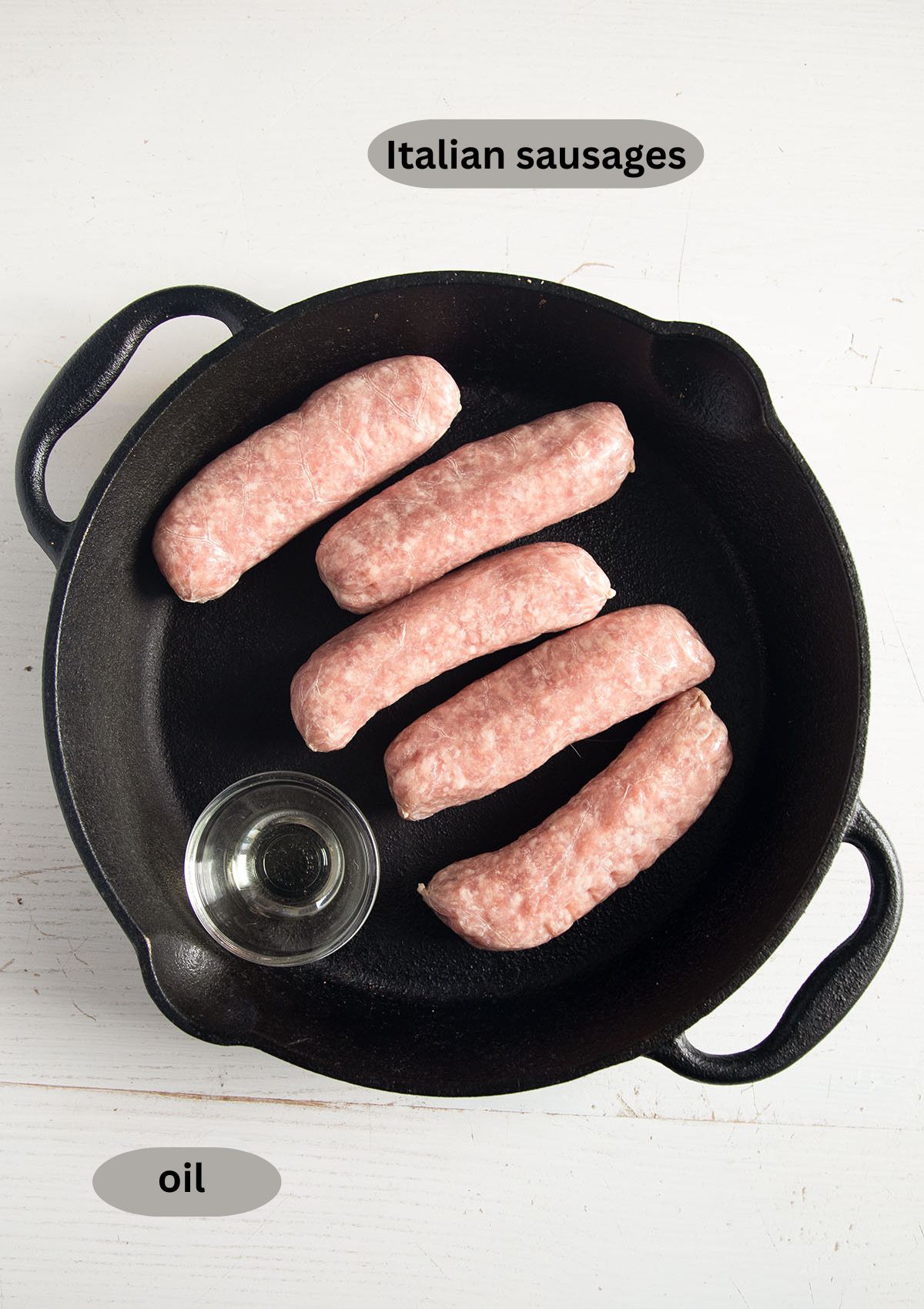 five italian sausage links and a small bowl with oil in a cast iron skillet, both labeled.