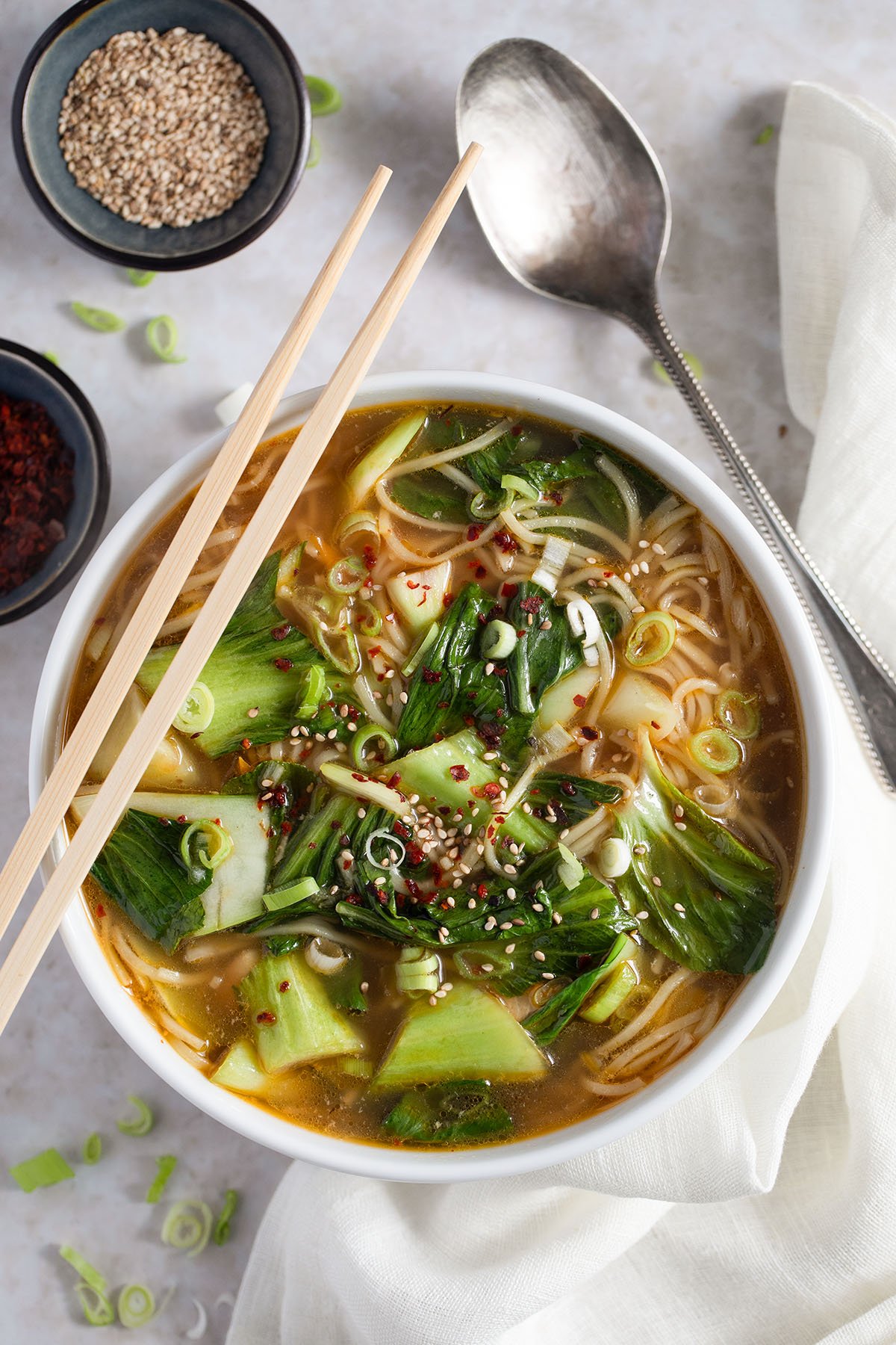 asian-inspired soup with chicken breast, pak choi, noodles and carrots in a bowl, small bowls with sesame and soy sauce and a spoon around it.