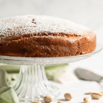 pistachio ricotta cake on a tall cake platter and some pistachios with shell scatter around it.
