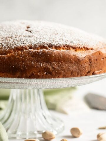 pistachio ricotta cake on a tall cake platter and some pistachios with shell scatter around it.