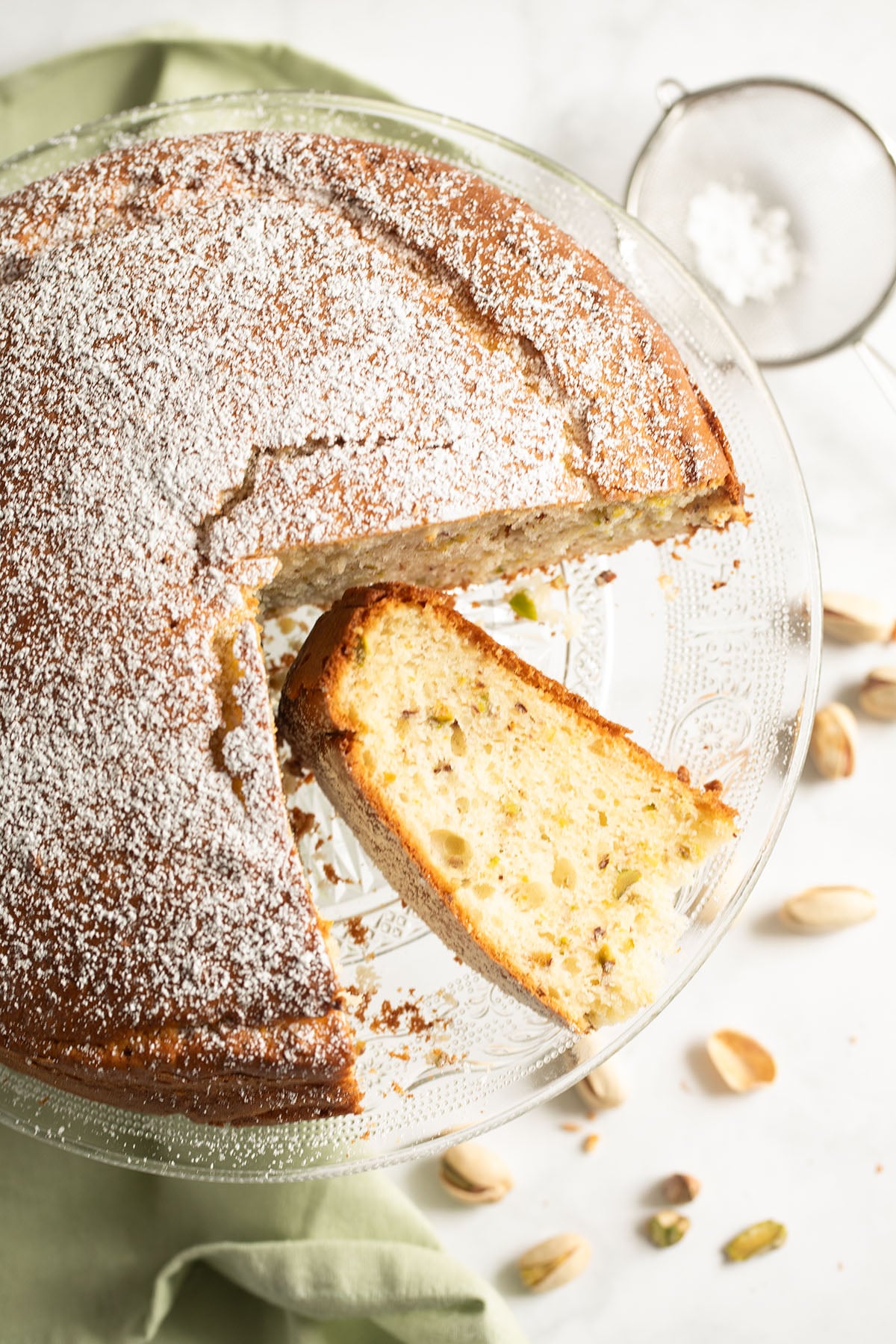 pistachio cake with ricotta with a slice showing the crumb on a platter with pistachios scattered around it.