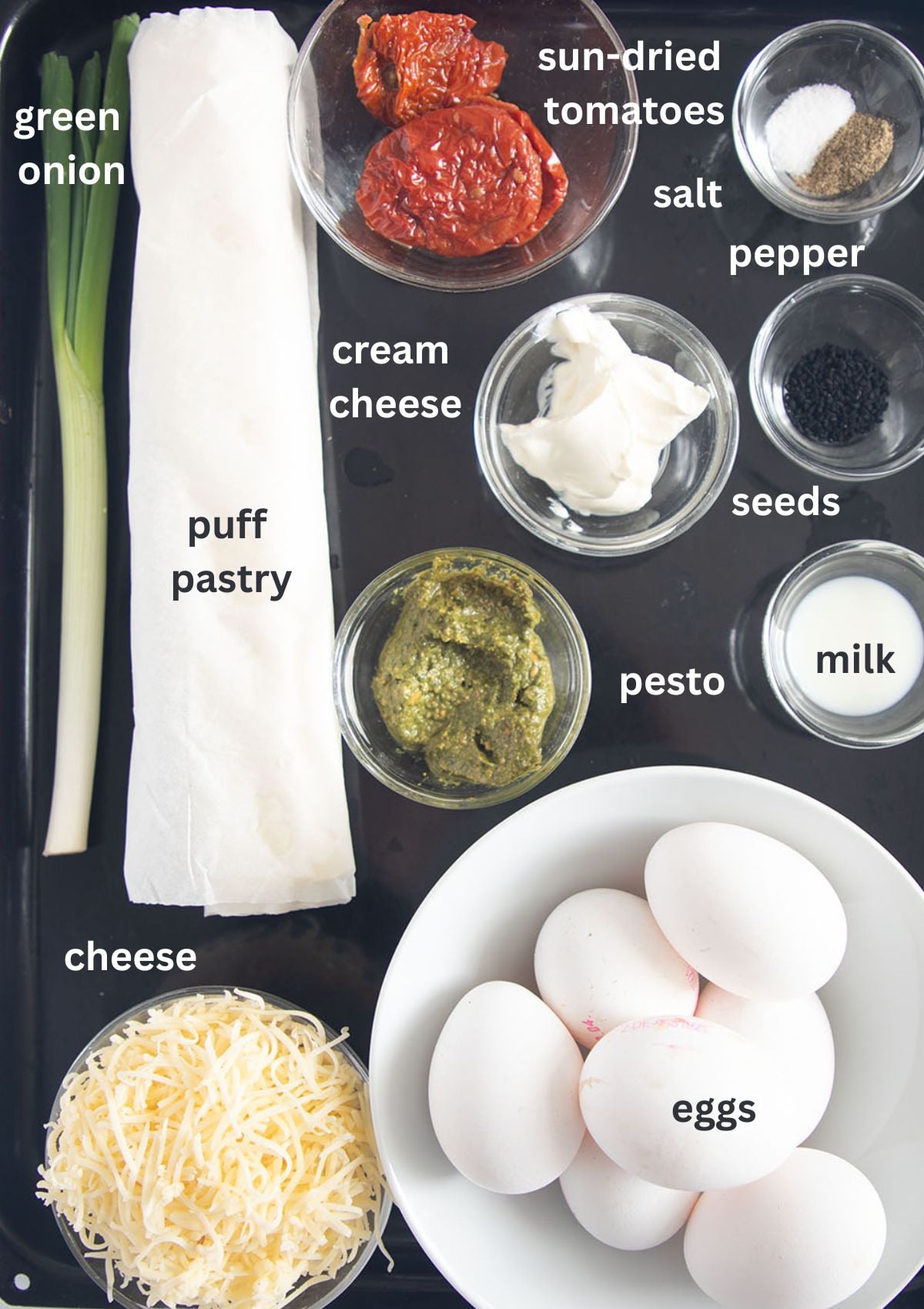 ingredients for making tarts with puff pastry, eggs, cheese, sun-dried tomatoes, pesto, cream cheese, and green onion.