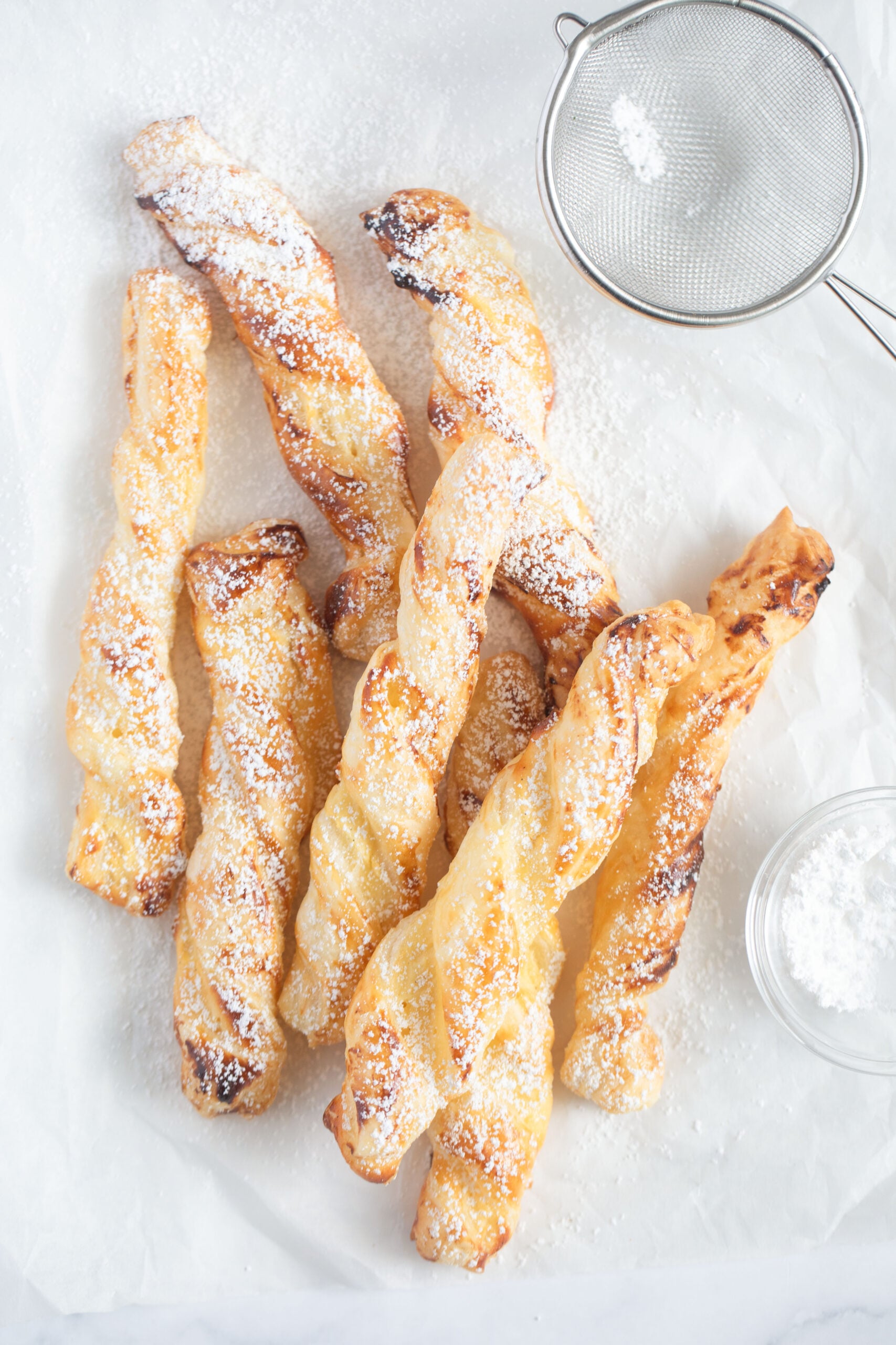 many puff pastry cream cheese twists, a small sieve and a bowl of icing sugar on parchment paper. 