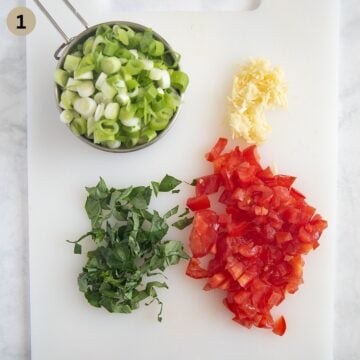 chopped green onions, chopped tomatoes, sliced basil leaves and grated garlic on a cutting board.