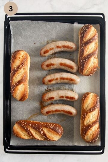 five bratwurst and 4 pretzel buns on a baking sheet.