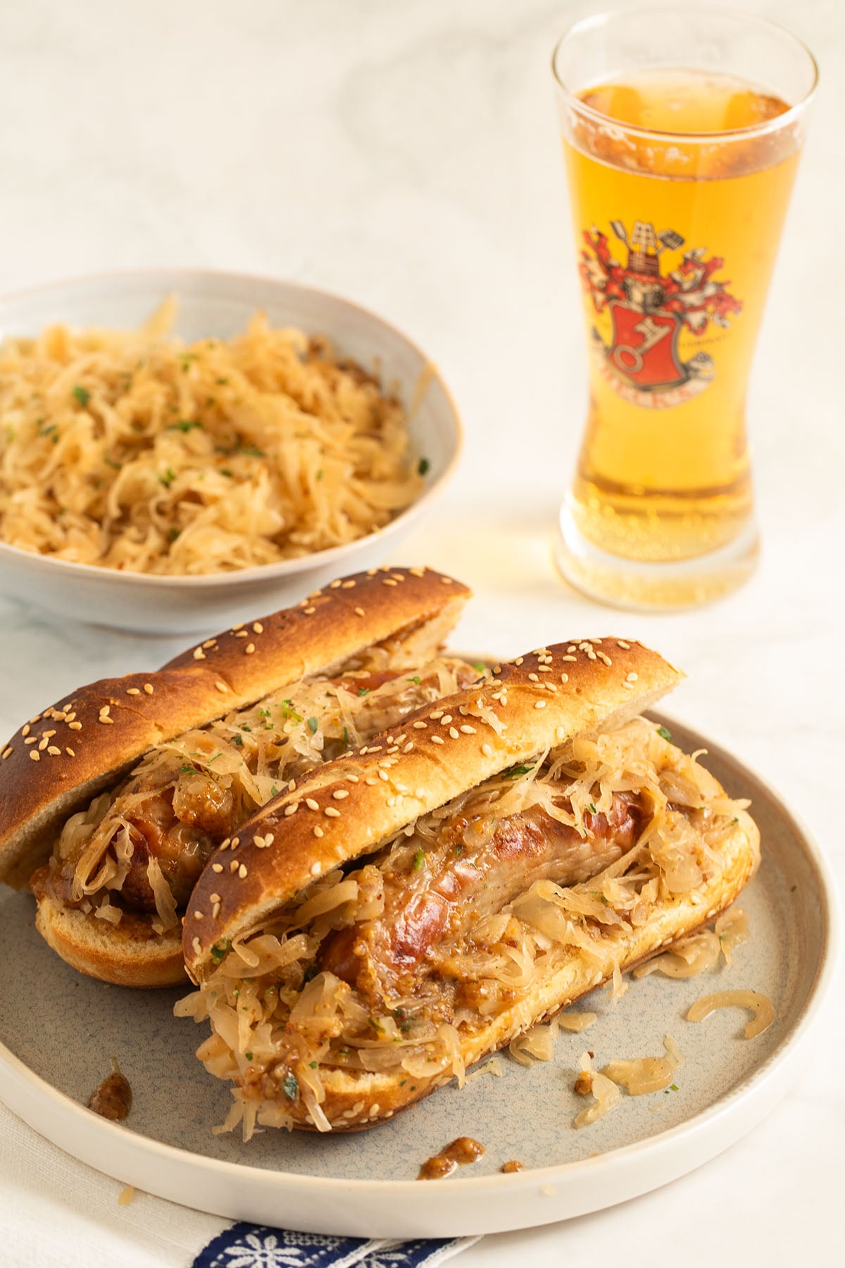 pretzel bun and bratwurst sandwiches on a plate, some sauerkraut and a glass of beer.