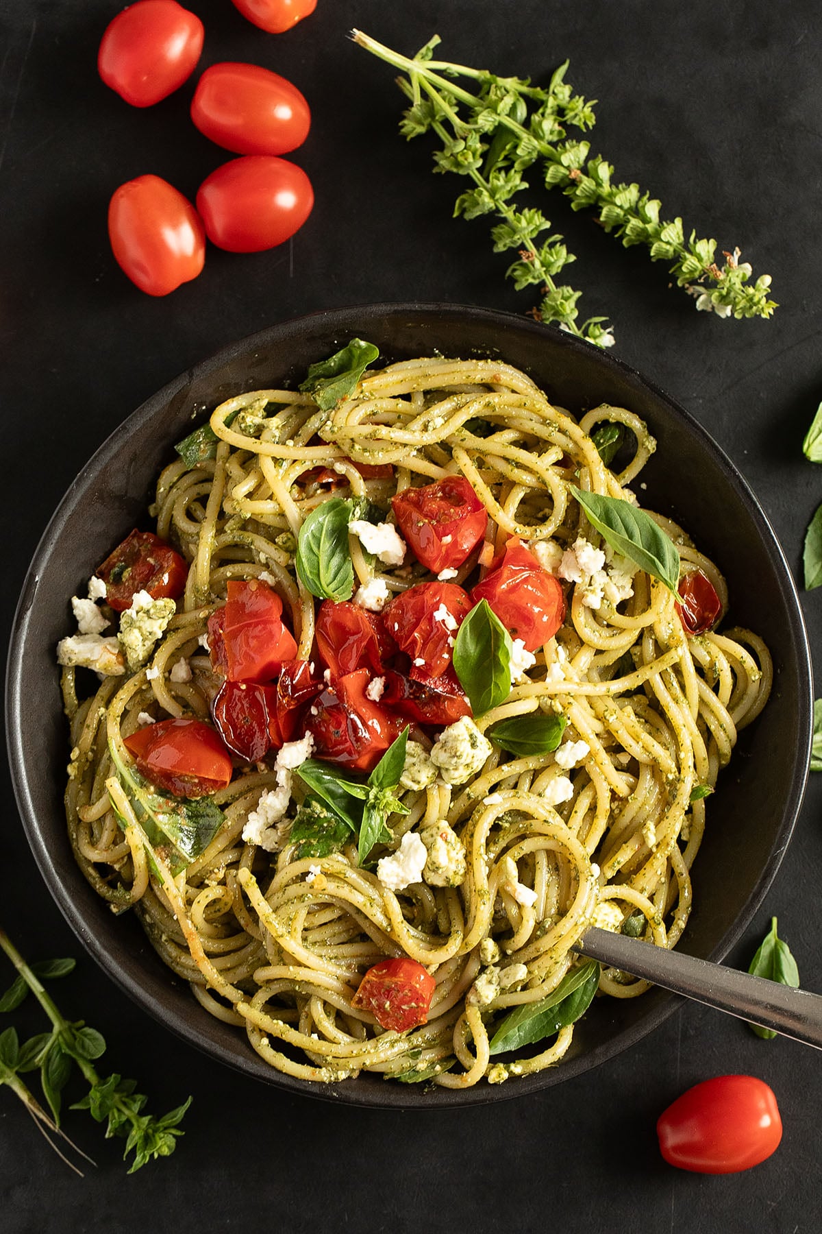 pasta with feta, pesto, roasted tomatoes, and basil in a bowl, more small tomatoes and fresh basil around it.