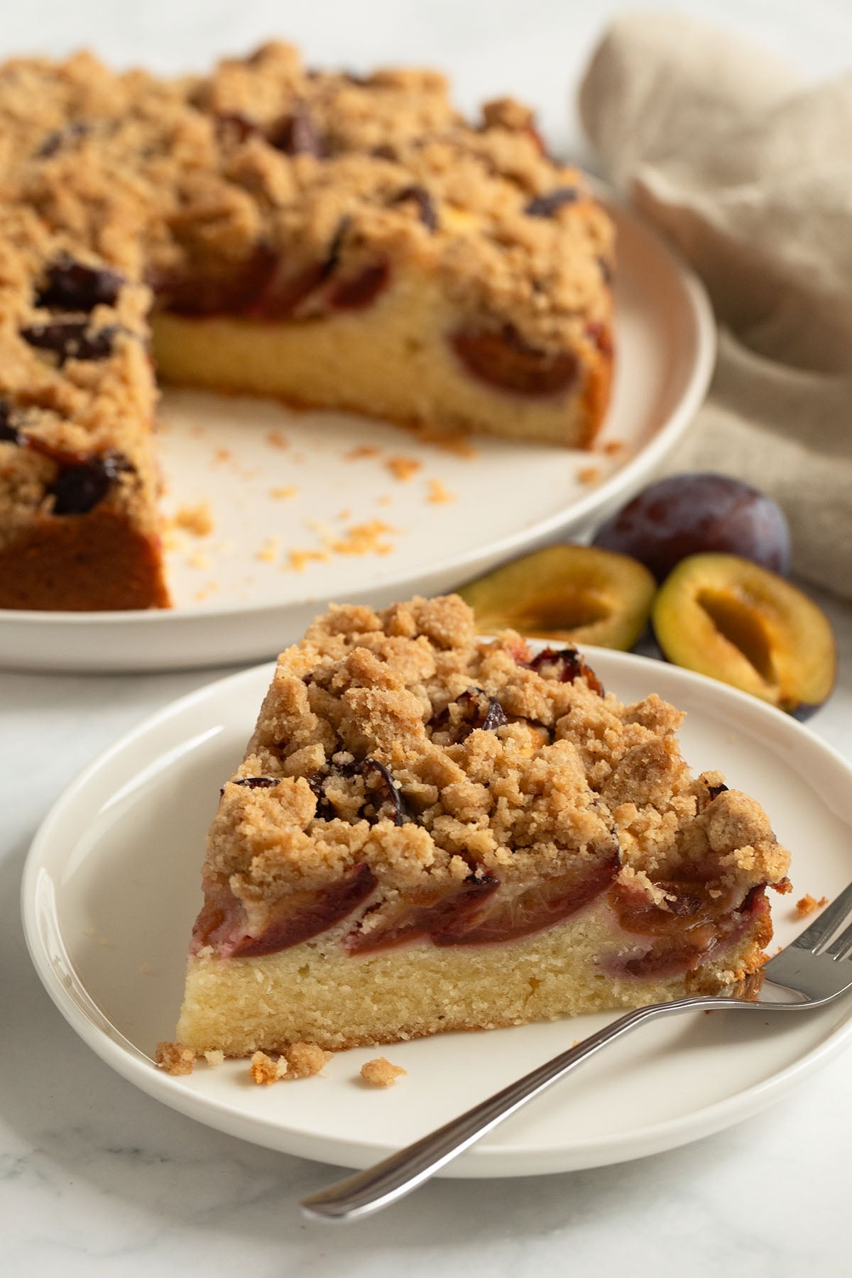 a thick slice of plum crumble cake on a plate and the rest of the cake behind.